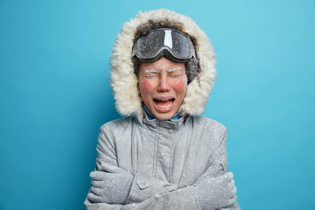 Photo of disappointed woman with red frozen face cries as feels very cold spent much time outdoor during severe frosty winter day wears grey outerwear trembles and cuddles herself. Recreation concept